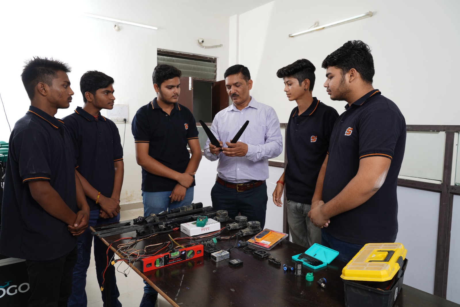 An instructor and four students in dark blue shirts gathered around a table with various drone components and tools during a technical training session.