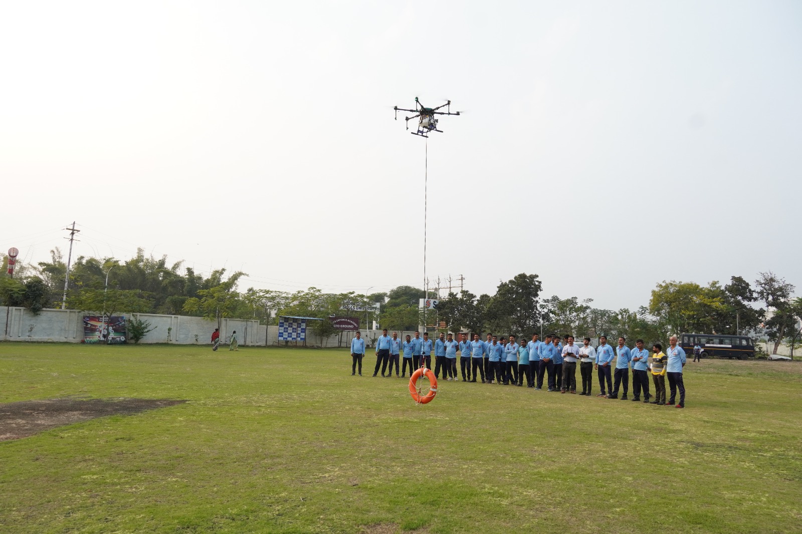 A group of trainees in light blue uniforms standing in a field watching a drone hover above them at the training ground.